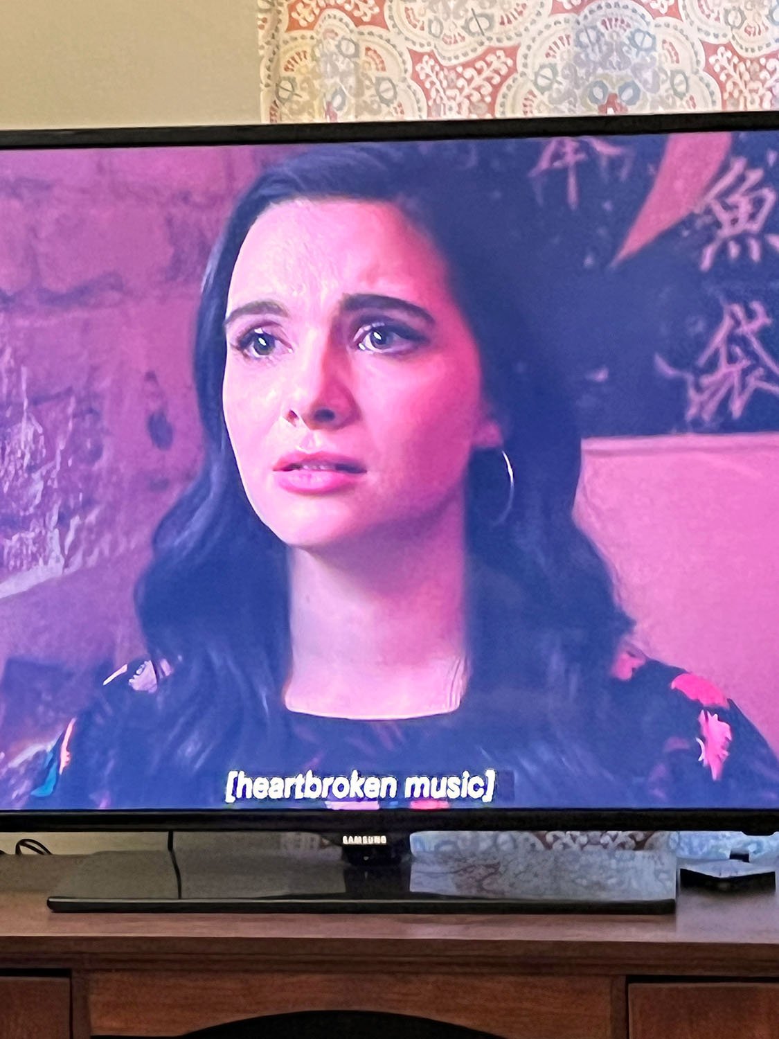 TV Screen with image of actress Katie Stevens, crying. Subtitle overlay reads, “heartbroken music.” 